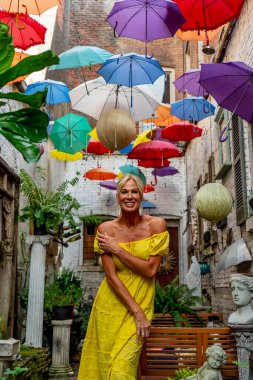 Güzel sarışın bir kadın zarif bir şekilde canlı ve renkli şemsiyelerin altında geziniyor. Neşeli ifadesi ve hafif adımları renkli sahnenin neşeli, canlı atmosferine ekleniyor..