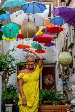 Güzel sarışın bir kadın zarif bir şekilde canlı ve renkli şemsiyelerin altında geziniyor. Neşeli ifadesi ve hafif adımları renkli sahnenin neşeli, canlı atmosferine ekleniyor..