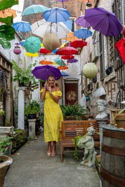 Güzel sarışın bir kadın zarif bir şekilde canlı ve renkli şemsiyelerin altında geziniyor. Neşeli ifadesi ve hafif adımları renkli sahnenin neşeli, canlı atmosferine ekleniyor..