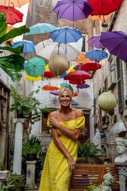 Güzel sarışın bir kadın zarif bir şekilde canlı ve renkli şemsiyelerin altında geziniyor. Neşeli ifadesi ve hafif adımları renkli sahnenin neşeli, canlı atmosferine ekleniyor..