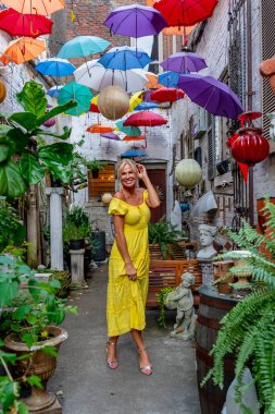 Güzel sarışın bir kadın zarif bir şekilde canlı ve renkli şemsiyelerin altında geziniyor. Neşeli ifadesi ve hafif adımları renkli sahnenin neşeli, canlı atmosferine ekleniyor..