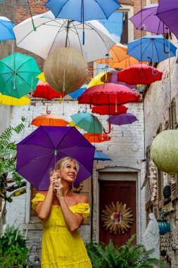 Güzel sarışın bir kadın zarif bir şekilde canlı ve renkli şemsiyelerin altında geziniyor. Neşeli ifadesi ve hafif adımları renkli sahnenin neşeli, canlı atmosferine ekleniyor..