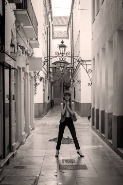 Çekici Avrupalı bir kadın hareketli bir İspanyol caddesinde kendine güvenen bir poz vererek, kıvrımlarını şık bir kıyafetle ortaya koyuyor. Güneşli arka plan ve hareketli atmosfer onun şık ve zahmetsiz zarafetini ön plana çıkarıyor..