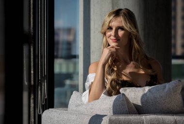 Çekici sarışın bir iç çamaşırı modeli rahat bir evde zarifçe poz veriyor, kıvrımları pencerelerden süzülen sıcak güneş ışığıyla vurgulanıyor, narin dantelli elbisesine yumuşak, altın bir parıltı saçıyor..