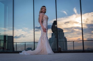 A beautiful blonde model, glowing in the sunlight, poses outdoors in a stunning wedding dress. The mirrored building behind her reflects the light, enhancing her radiance and creating a captivating scene. clipart