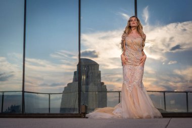 Güneşin altında parlayan güzel bir sarışın manken çarpıcı bir gelinlikle dışarıda poz veriyor. Arkasındaki aynalı bina ışığı yansıtıyor, ışığını arttırıyor ve büyüleyici bir sahne yaratıyor..