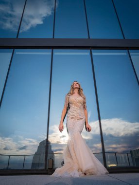 Güneşin altında parlayan güzel bir sarışın manken çarpıcı bir gelinlikle dışarıda poz veriyor. Arkasındaki aynalı bina ışığı yansıtıyor, ışığını arttırıyor ve büyüleyici bir sahne yaratıyor..