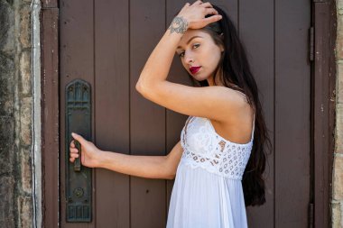 Çekici genç bir esmer, dalgalı beyaz bir bahar elbisesi içinde, saçları dalgalar halinde dalgalanırken dışarıda poz veriyor. Gülümsüyor, uzun ve sert bir kışın ardından baharın yumuşak sıcağında güneşleniyor..