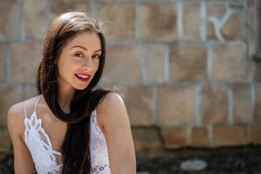 Çekici genç bir esmer, dalgalı beyaz bir bahar elbisesi içinde, saçları dalgalar halinde dalgalanırken dışarıda poz veriyor. Gülümsüyor, uzun ve sert bir kışın ardından baharın yumuşak sıcağında güneşleniyor..