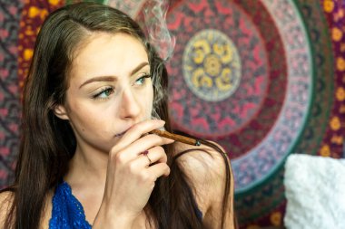 A gorgeous young brunette relaxes in her private home, smoking marijuana. She takes a slow inhale, using it as a calming escape to help manage the stresses and challenges of her daily life. clipart