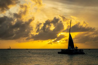 Parlak altın bir günbatımı, sakin sularda parıldayan bir ışık yayar. Siluetli yelkenliler usulca sürükleniyorlar. Karanlık şekilleri gökyüzünün ateşli renkleriyle çelişiyor. Huzur ve merakı çağrıştırıyor..