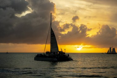 Parlak altın bir günbatımı, sakin sularda parıldayan bir ışık yayar. Siluetli yelkenliler usulca sürükleniyorlar. Karanlık şekilleri gökyüzünün ateşli renkleriyle çelişiyor. Huzur ve merakı çağrıştırıyor..