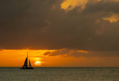 Parlak altın bir günbatımı, sakin sularda parıldayan bir ışık yayar. Siluetli yelkenliler usulca sürükleniyorlar. Karanlık şekilleri gökyüzünün ateşli renkleriyle çelişiyor. Huzur ve merakı çağrıştırıyor..