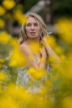 Güzel bir sarışın model, canlı sarı çiçeklerle dolu bir alanda geziniyor, portreler için çarpıcı pozlar veriyor. Yaz sonu güneşi sahnenin üzerine sıcacık bir parıltı saçıyor, zarafetini ve doğal güzelliğini vurguluyor..