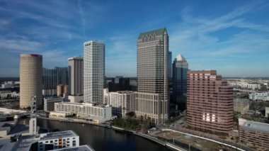 Tampa, Florida 'nın hava manzarası modern yüksek binalarla dolu canlı bir ufuk çizgisi, şehir merkezinden geçen ikonik Hillsborough Nehri, Tampa Körfezi boyunca geniş deniz kenarları ve yemyeşil noktalı kentsel alanlar gösteriyor..