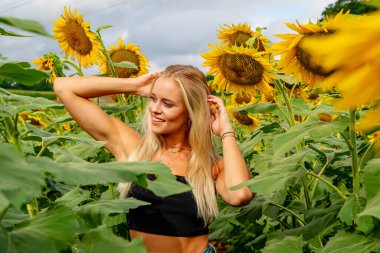 Çekici sarışın bir model canlı bir ayçiçeği tarlasında zarifçe poz veriyor, yaz gününün sıcağında güneşleniyor. Altın çiçekler etrafını sarmış, neşeli ve göz alıcı bir açık hava manzarası yaratıyor..