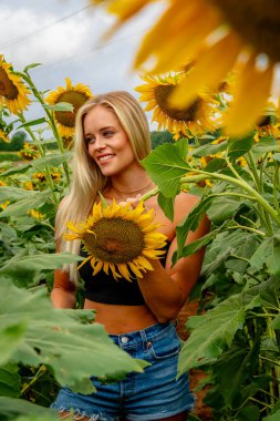 Çekici sarışın bir model canlı bir ayçiçeği tarlasında zarifçe poz veriyor, yaz gününün sıcağında güneşleniyor. Altın çiçekler etrafını sarmış, neşeli ve göz alıcı bir açık hava manzarası yaratıyor..