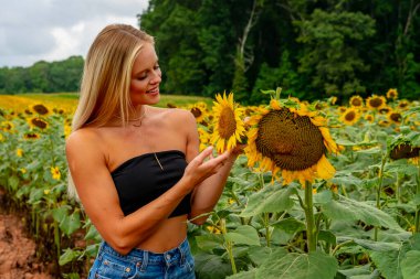 Çekici sarışın bir model canlı bir ayçiçeği tarlasında zarifçe poz veriyor, yaz gününün sıcağında güneşleniyor. Altın çiçekler etrafını sarmış, neşeli ve göz alıcı bir açık hava manzarası yaratıyor..