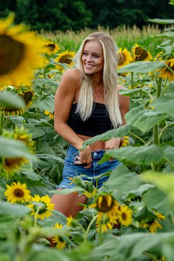 Çekici sarışın bir model canlı bir ayçiçeği tarlasında zarifçe poz veriyor, yaz gününün sıcağında güneşleniyor. Altın çiçekler etrafını sarmış, neşeli ve göz alıcı bir açık hava manzarası yaratıyor..