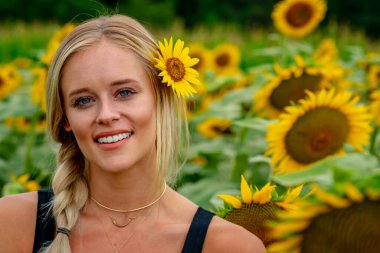 Çekici sarışın bir model canlı bir ayçiçeği tarlasında zarifçe poz veriyor, yaz gününün sıcağında güneşleniyor. Altın çiçekler etrafını sarmış, neşeli ve göz alıcı bir açık hava manzarası yaratıyor..