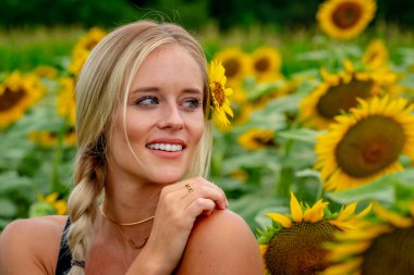 Çekici sarışın bir model canlı bir ayçiçeği tarlasında zarifçe poz veriyor, yaz gününün sıcağında güneşleniyor. Altın çiçekler etrafını sarmış, neşeli ve göz alıcı bir açık hava manzarası yaratıyor..