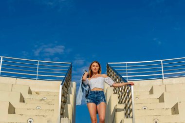 Çekici genç bir sarışın model şehir merkezinde poz veriyor. Şık yaz kıyafeti parlak mavi gökyüzüne tamamen zıt düşüyor. Kendine güveni tam, sıcaklığı ve zahmetsizliği yayıyor..