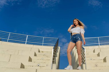 Çekici genç bir sarışın model şehir merkezinde poz veriyor. Şık yaz kıyafeti parlak mavi gökyüzüne tamamen zıt düşüyor. Kendine güveni tam, sıcaklığı ve zahmetsizliği yayıyor..