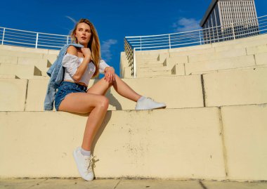 Çekici genç bir sarışın model şehir merkezinde poz veriyor. Şık yaz kıyafeti parlak mavi gökyüzüne tamamen zıt düşüyor. Kendine güveni tam, sıcaklığı ve zahmetsizliği yayıyor..