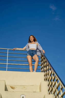 Çekici genç bir sarışın model şehir merkezinde poz veriyor. Şık yaz kıyafeti parlak mavi gökyüzüne tamamen zıt düşüyor. Kendine güveni tam, sıcaklığı ve zahmetsizliği yayıyor..