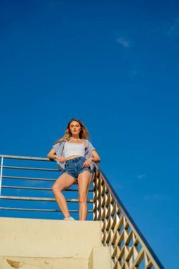 Çekici genç bir sarışın model şehir merkezinde poz veriyor. Şık yaz kıyafeti parlak mavi gökyüzüne tamamen zıt düşüyor. Kendine güveni tam, sıcaklığı ve zahmetsizliği yayıyor..