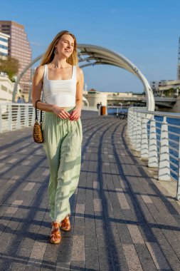 A stunning young blonde model strikes a pose downtown, her chic summer outfit perfectly contrasting against the vibrant blue sky. She exudes confidence, radiating warmth and effortless style. clipart