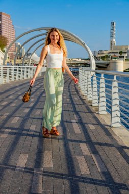 Çekici genç bir sarışın model şehir merkezinde poz veriyor. Şık yaz kıyafeti parlak mavi gökyüzüne tamamen zıt düşüyor. Kendine güveni tam, sıcaklığı ve zahmetsizliği yayıyor..