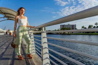 Çekici genç bir sarışın model şehir merkezinde poz veriyor. Şık yaz kıyafeti parlak mavi gökyüzüne tamamen zıt düşüyor. Kendine güveni tam, sıcaklığı ve zahmetsizliği yayıyor..