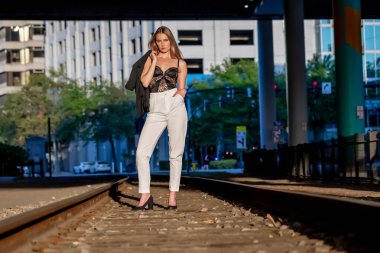 Çekici genç sarışın bir kadın zarif bir şekilde poz verir, batan güneşin sıcacık parıltısıyla yıkanır. Şehir arkaplanı onun tarzını geliştirir. Güzellik ve şehir cazibesinin büyüleyici bir karışımını yaratır..