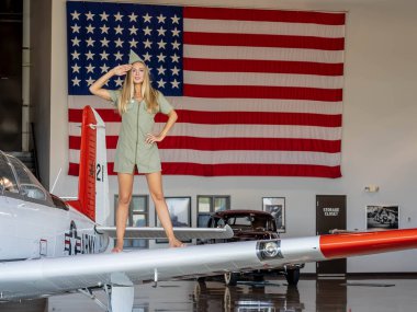 Çarpıcı sarışın bir model, dev bir Amerikan bayrağıyla çerçevelenmiş, klasik Americana kıyafetleriyle, eski bir 2. Dünya Savaşı uçağının yanında poz veriyor. Amerikan Hava Kuvvetleri 'nin geri kalmış görüntüsü vatansever bir tarz yayıyor..