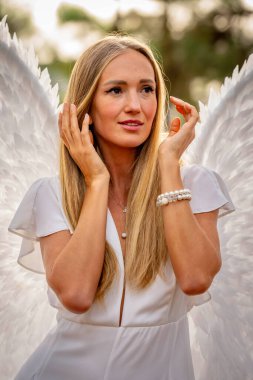 A stunning young blonde dressed in pure white, adorned with delicate angel wings, strikes a graceful pose. Her serene expression and flowing attire evoke an ethereal presence, embodying angelic beauty. clipart