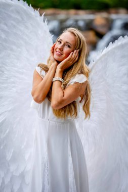 A stunning young blonde dressed in pure white, adorned with delicate angel wings, strikes a graceful pose. Her serene expression and flowing attire evoke an ethereal presence, embodying angelic beauty. clipart