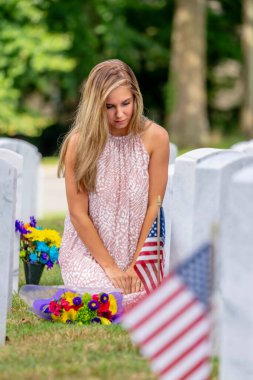 A young bride kneels at a military cemetery amid uniform rows of graves. Clutching flowers and a tombstone, tears streak her face as she mourns a beloved family member with profound, silent grief. clipart