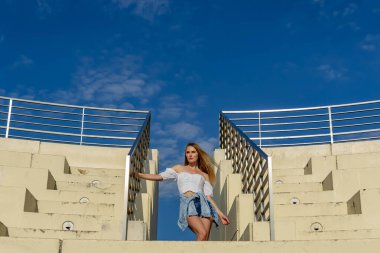 Çekici genç bir sarışın model şehir merkezinde poz veriyor. Şık yaz kıyafeti parlak mavi gökyüzüne tamamen zıt düşüyor. Kendine güveni tam, sıcaklığı ve zahmetsizliği yayıyor..