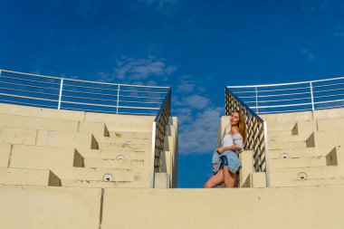 Çekici genç bir sarışın model şehir merkezinde poz veriyor. Şık yaz kıyafeti parlak mavi gökyüzüne tamamen zıt düşüyor. Kendine güveni tam, sıcaklığı ve zahmetsizliği yayıyor..