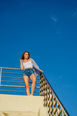 Çekici genç bir sarışın model şehir merkezinde poz veriyor. Şık yaz kıyafeti parlak mavi gökyüzüne tamamen zıt düşüyor. Kendine güveni tam, sıcaklığı ve zahmetsizliği yayıyor..