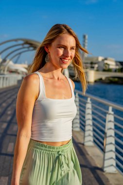 Çekici genç bir sarışın model şehir merkezinde poz veriyor. Şık yaz kıyafeti parlak mavi gökyüzüne tamamen zıt düşüyor. Kendine güveni tam, sıcaklığı ve zahmetsizliği yayıyor..