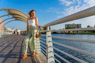 Çekici genç bir sarışın model şehir merkezinde poz veriyor. Şık yaz kıyafeti parlak mavi gökyüzüne tamamen zıt düşüyor. Kendine güveni tam, sıcaklığı ve zahmetsizliği yayıyor..