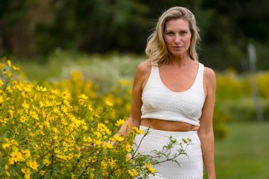 Güzel bir sarışın model, canlı sarı çiçeklerle dolu bir alanda geziniyor, portreler için çarpıcı pozlar veriyor. Yaz sonu güneşi sahnenin üzerine sıcacık bir parıltı saçıyor, zarafetini ve doğal güzelliğini vurguluyor..