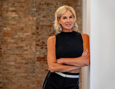 Güzel sarışın bir kadın kendine güvenerek ayakta duruyor, ışıl ışıl gülüşü ve kendine güvenen tavırları zarafet saçıyor. Zarif duruşu çekiciliğini vurguluyor, zamansız güzellik ve kendine güvenin görüntüsünü yaratıyor..