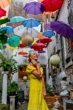 Güzel sarışın bir kadın zarif bir şekilde canlı ve renkli şemsiyelerin altında geziniyor. Neşeli ifadesi ve hafif adımları renkli sahnenin neşeli, canlı atmosferine ekleniyor..