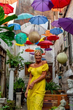 Güzel sarışın bir kadın zarif bir şekilde canlı ve renkli şemsiyelerin altında geziniyor. Neşeli ifadesi ve hafif adımları renkli sahnenin neşeli, canlı atmosferine ekleniyor..
