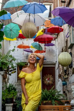 Güzel sarışın bir kadın zarif bir şekilde canlı ve renkli şemsiyelerin altında geziniyor. Neşeli ifadesi ve hafif adımları renkli sahnenin neşeli, canlı atmosferine ekleniyor..