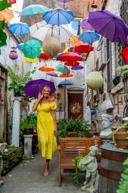 Güzel sarışın bir kadın zarif bir şekilde canlı ve renkli şemsiyelerin altında geziniyor. Neşeli ifadesi ve hafif adımları renkli sahnenin neşeli, canlı atmosferine ekleniyor..