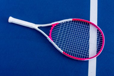 Tek bir tenis raketi güneş ışığıyla kaplı kortta duruyor, sapı taban çizgisiyle aynı hizada, kibritler arasında sakin bir anda mavi yüzey boyunca yumuşak bir gölge bırakıyor..